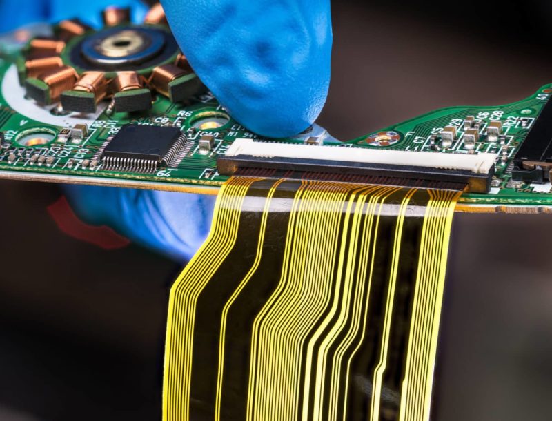 Flat plastic strip flexible cable on printed circuit board on a dark background Hand in blue glove holding green PCB with integrated motor stator and electronic components as micro chip or resistors. Surface mount technology